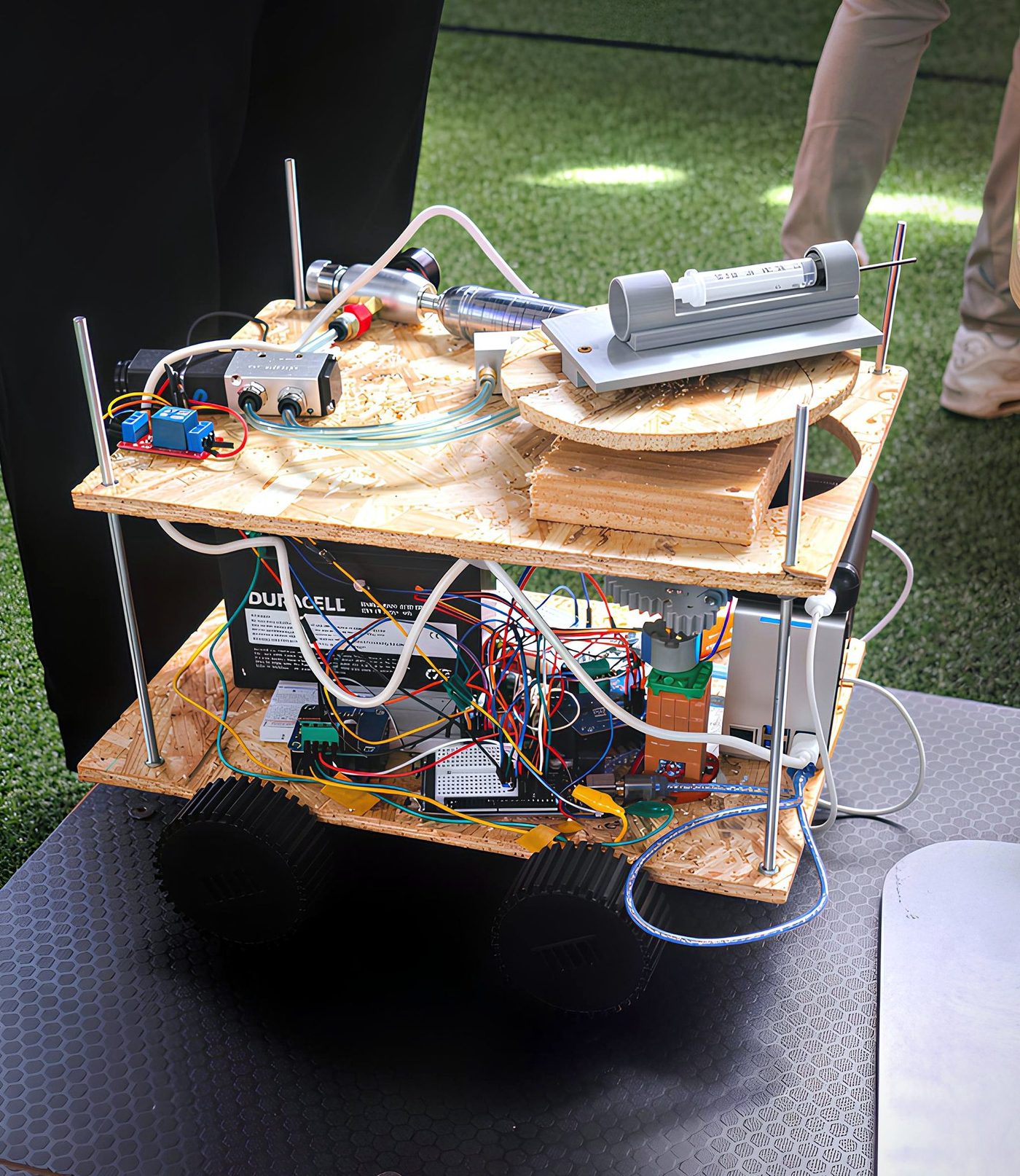 The Lynra rover: a two-tier plywood chassis carrying a battery, control electronics, a Raspberry Pi, pneumatics and a 3D-printed syringe assembly, on four treaded wheels.